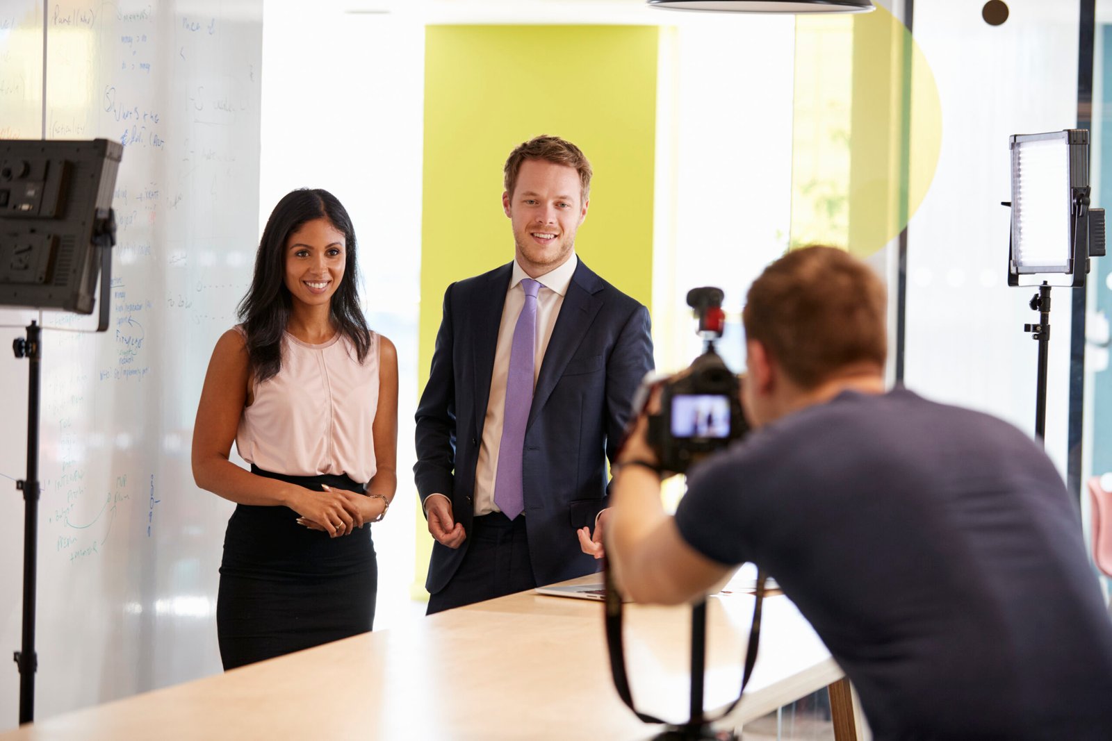 Photograph of two professionally dressed individuals, a woman and a man, standing in front of a camera setup in a modern office. A videographer is filming them, and studio lights are positioned on both sides. The background features a whiteboard with notes and a vibrant yellow-green wall, suggesting a corporate video shoot or promotional content creation.