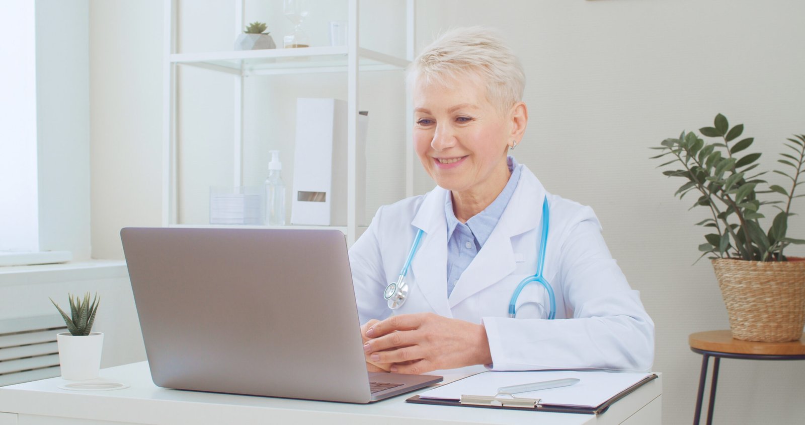 Senior female doctor smiling during a video call on a laptop in a bright medical office, symbolizing digital communication, patient engagement, and online reputation management in healthcare.